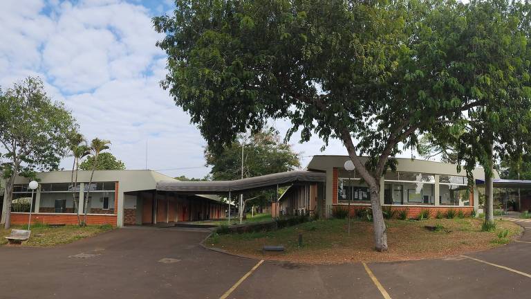 Imagem panorâmica de um edifício de um andar, cercado por árvores grandes e um céu parcialmente nublado. O edifício possui uma estrutura de telhado plano e janelas amplas. A área em frente ao edifício é pavimentada, com algumas linhas de estacionamento visíveis.