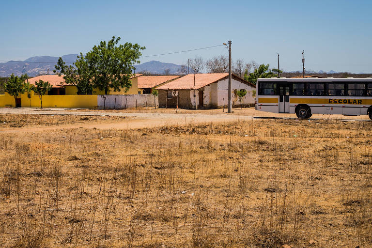 A imagem mostra um cenário rural com um ônibus escolar estacionado em uma área seca. Ao fundo, há algumas casas com telhados vermelhos e uma parede amarela. Árvores estão visíveis à esquerda, e montanhas podem ser vistas ao longe sob um céu claro.