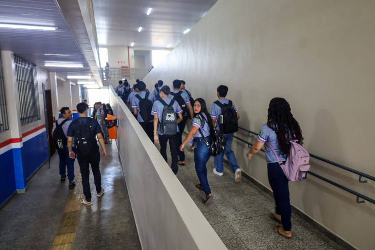 Um grupo de estudantes caminha por um corredor largo e iluminado, com paredes claras. Eles estão usando mochilas e vestindo camisetas azuis. O corredor possui uma rampa acessível à direita e é decorado com uma faixa colorida na parede. Ao fundo, há portas e janelas.