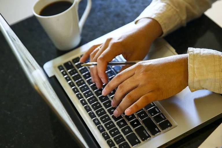 A imagem mostra uma pessoa digitando em um laptop. A mão direita está segurando uma caneta, enquanto a mão esquerda está sobre o teclado. Ao lado do laptop, há uma xícara de café e um par de óculos sobre uma folha de papel. O ambiente parece ser uma mesa de trabalho com superfície escura.