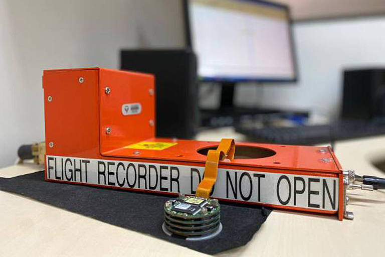 A imagem mostra um gravador de voo, também conhecido como caixa-preta, em uma mesa. O dispositivo é de cor laranja e possui uma etiqueta que diz 'FLIGHT RECORDER DO NOT OPEN'. Ao fundo, há um computador com uma tela visível.