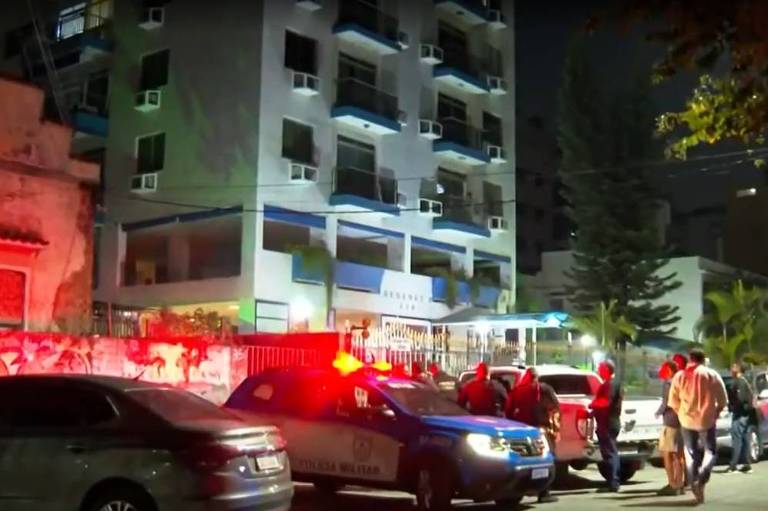 A imagem mostra um prédio de apartamentos à noite, com várias janelas iluminadas e ar-condicionado visível. Em frente ao prédio, há um grupo de pessoas e um carro da polícia com luzes vermelhas piscando. O ambiente é urbano, com árvores ao fundo e outros veículos estacionados nas proximidades.