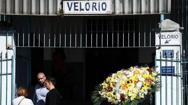 A imagem mostra um local com uma placa escrito "velório", com uma coroa de flores do lado direito e três pessoas conversando do lado esquerdo