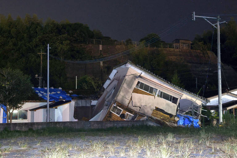 A imagem mostra uma casa inclinada, aparentemente danificada por um terremoto, com a estrutura se apoiando em um ângulo instável. Ao fundo, há uma vegetação densa e uma elevação. A cena é noturna, com iluminação artificial visível. Outras construções estão presentes, algumas cobertas com lonas azuis.