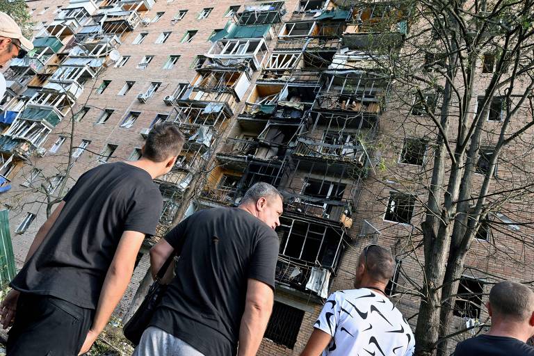 A imagem mostra um grupo de pessoas observando um edifício danificado. O prédio, de vários andares, apresenta janelas quebradas e varandas em estado precário. Algumas partes da fachada estão expostas, revelando danos significativos. As pessoas estão em primeiro plano, voltadas para o edifício, e parecem estar discutindo a situação.