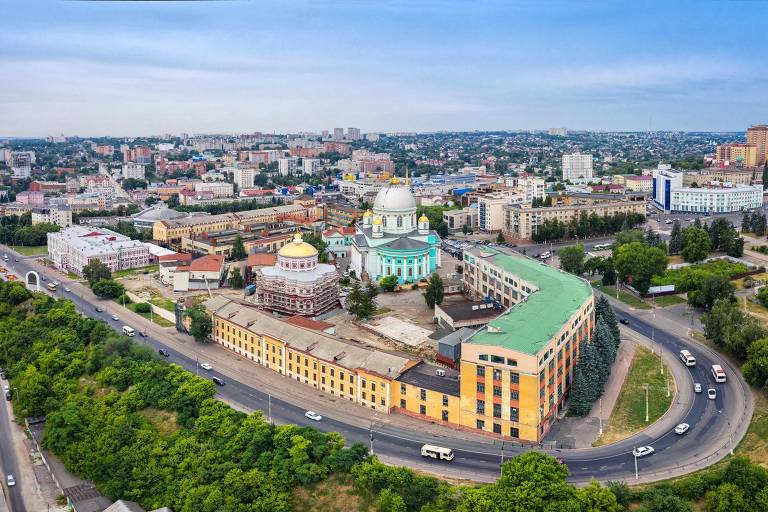 A imagem mostra uma vista aérea de uma cidade com diversos edifícios e uma estrada em primeiro plano. No centro, destaca-se um grande edifício com cúpulas coloridas, incluindo uma cúpula verde e outra dourada. Ao redor, há várias construções de diferentes estilos e tamanhos, além de áreas verdes. A cidade parece ser bastante urbanizada, com veículos circulando nas estradas.