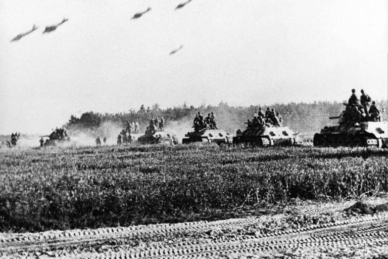 A imagem mostra uma formação de tanques em um campo, com soldados visíveis em cima deles. O cenário é em preto e branco, e há uma nuvem de poeira levantada pelo movimento dos veículos. No céu, é possível ver vários aviões voando. O ambiente parece ser rural, com árvores ao fundo.