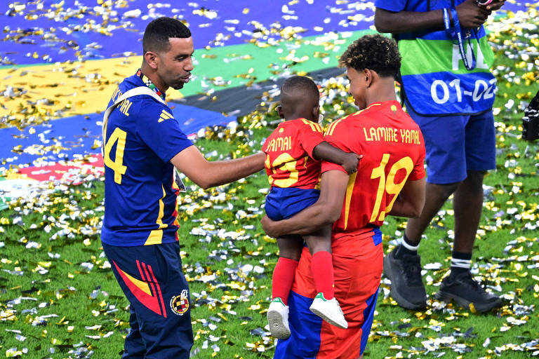 A imagem mostra três pessoas em um campo de futebol coberto de confetes. Um homem, vestido com uma camisa azul e calças escuras, está conversando com um menino pequeno que usa uma camiseta vermelha com o nome 'Yamal' nas costas. Outro jovem, que segura o menino, está vestido com uma camiseta vermelha e calças que combinam, com o nome 'Lamine Yamal' nas costas. Ao fundo, há um homem com uma câmera e confetes espalhados pelo chão.
