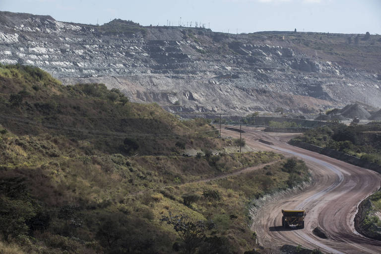 A imagem mostra uma estrada sinuosa em uma área de mineração, com um grande caminhão de carga se deslocando. Ao fundo, há uma encosta de terra com camadas expostas, indicando a atividade de extração mineral. A vegetação é escassa nas proximidades da estrada, e o céu está parcialmente nublado.
