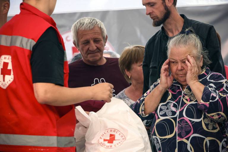 Homem de jaleco vermelho com uma cruz atrás dá uma sacola a uma senhora de cabelo grisalho com vestido arroxeado, com as mãos à cabeça. Outros dois homens, um alto com barba escura e outro idoso de cabelo branco, além de um menino de cabelo loiro, compõem a cena