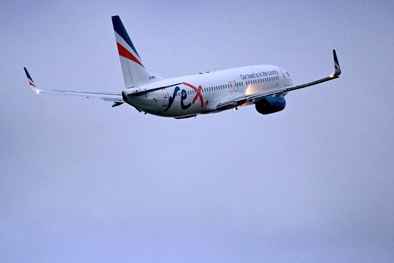 A imagem mostra um avião comercial em voo, inclinado para a direita, com as asas estendidas e a cauda visível. O céu ao fundo é nublado, em tons de azul e cinza.