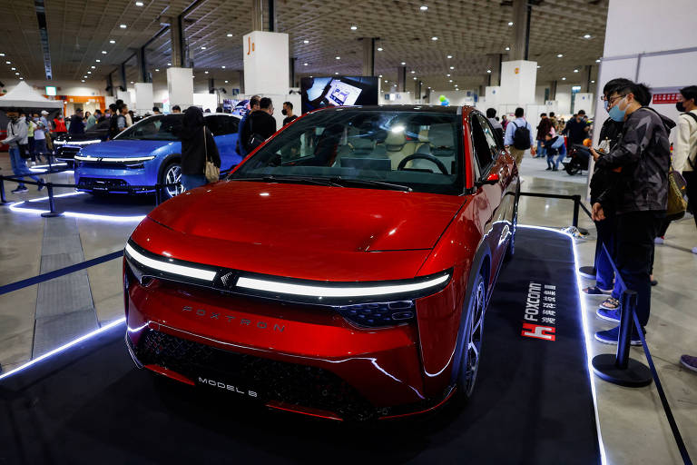 A imagem mostra um carro vermelho em uma exposição, com um design moderno e futurista. O veículo está em destaque, cercado por uma iluminação que realça suas linhas. Ao fundo, há outras pessoas e um carro azul, indicando um evento de automóveis. O ambiente parece ser um grande espaço de exposição, com várias pessoas observando os veículos.