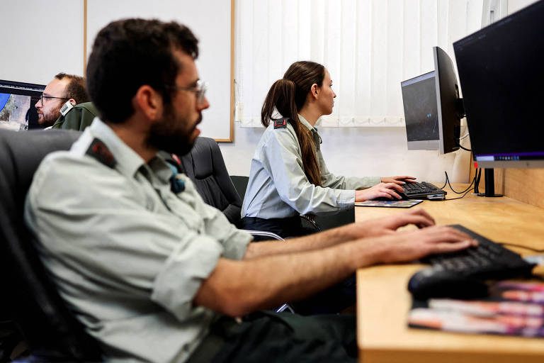 A imagem mostra três pessoas em um ambiente de escritório. Duas delas estão sentadas em cadeiras, utilizando computadores. Um homem, à esquerda, está focado na tela do computador, enquanto uma mulher, à direita, também está concentrada em seu trabalho. Ao fundo, há outra pessoa visível, parcialmente, em uma mesa. O ambiente é bem iluminado e organizado.