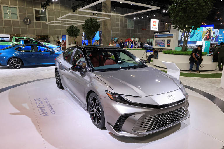 A imagem mostra um carro modelo Toyota Camry 2023, de cor prata, em um estande de exposição. O veículo está posicionado em um pedestal circular com o texto 'THE ALL-ELECTRIFIED 2023 CAMRY' visível. Ao fundo, há outros carros, incluindo um modelo azul, e várias pessoas interagindo com os expositores. O ambiente é bem iluminado, com árvores decorativas e painéis de exibição.
