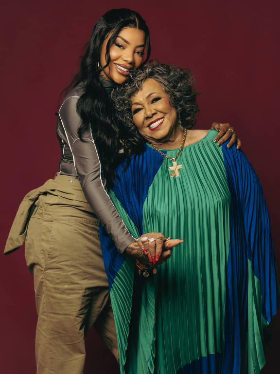 A imagem mostra duas mulheres sorrindo e posando juntas. A mulher à esquerda é mais jovem, com cabelo longo e liso, vestindo uma blusa cinza e calças claras. A mulher à direita é mais velha, com cabelo grisalho e encaracolado, usando um vestido plissado em tons de verde e azul. Ambas estão segurando as mãos uma da outra, com expressões alegres, contra um fundo vermelho.