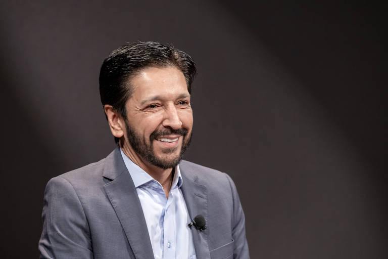 Um homem com cabelo escuro e barba， vestindo um terno cinza e uma camisa branca， está sorrindo enquanto é entrevistado. O fundo é escuro， destacando sua expressão amigável.