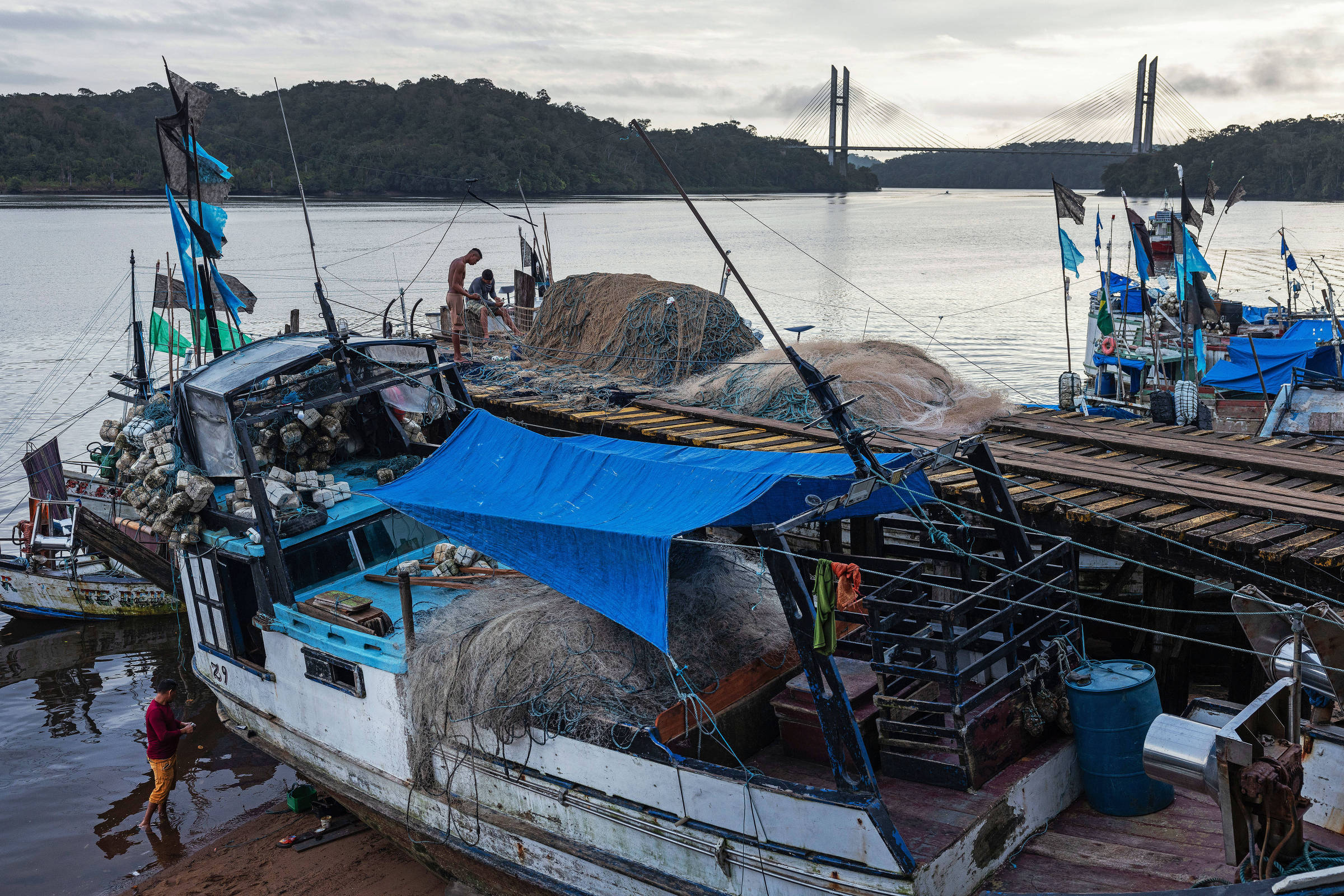 Em um píer de madeira homem segura rede com as mãos ao lado de dois barcos atracados com uma ponte estaiada ao fundo