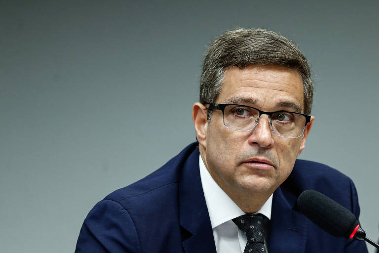 Um homem com cabelo curto e grisalho, usando óculos e um terno escuro, está sentado em uma mesa. Ele parece estar em uma conferência ou reunião, olhando para frente com uma expressão séria. O fundo é de uma cor cinza neutra.
