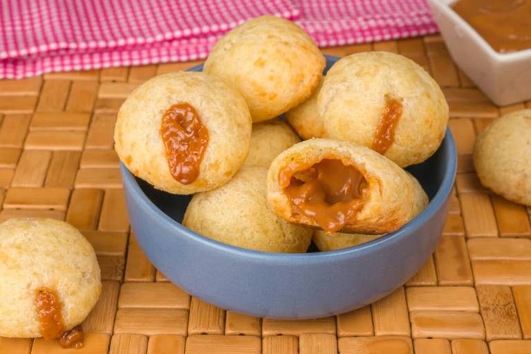 A imagem mostra alguns pães de queijo recheados com doce de leite dentro de um pote azul. Em volta deles se encontram alguns pães de queijo e um pote pequeno com doce de leite dentro