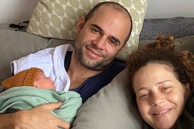 Em foto colorida, casal aparece com o filho em uma foto de família