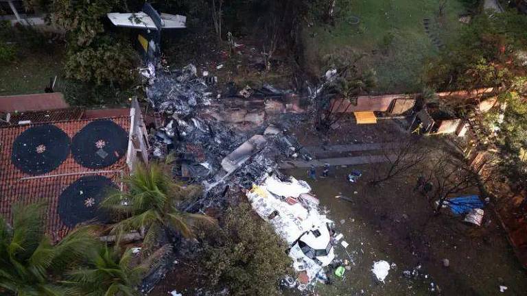 Imagem aérea de um local de acidente aéreo, mostrando destroços de uma aeronave espalhados em uma área com vegetação. Há partes da fuselagem e outros materiais de construção ao redor, com algumas árvores e uma estrutura de edifício visíveis ao fundo.