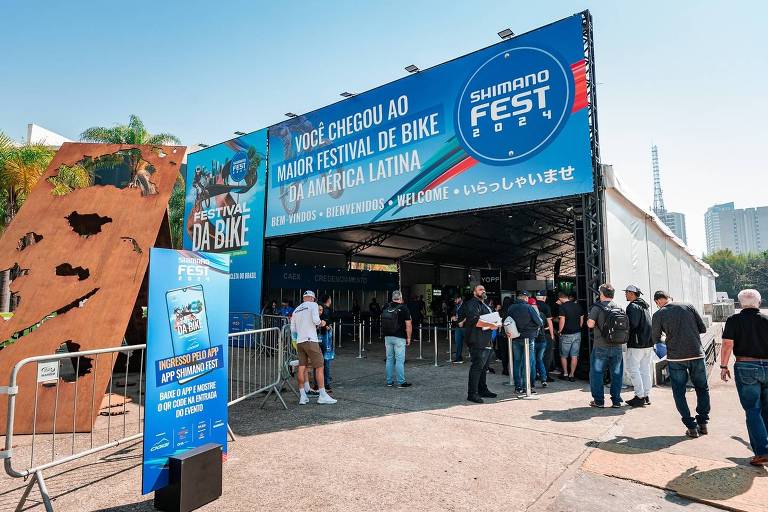 A imagem mostra a entrada de um evento chamado Gamer Fest. Há uma grande estrutura coberta com um banner azul que exibe o nome do evento e informações sobre a programação. Na frente, várias pessoas estão entrando, algumas em fila. À esquerda, há uma parede de escalada e, ao fundo, pode-se ver a cidade com prédios. O céu está claro e ensolarado.