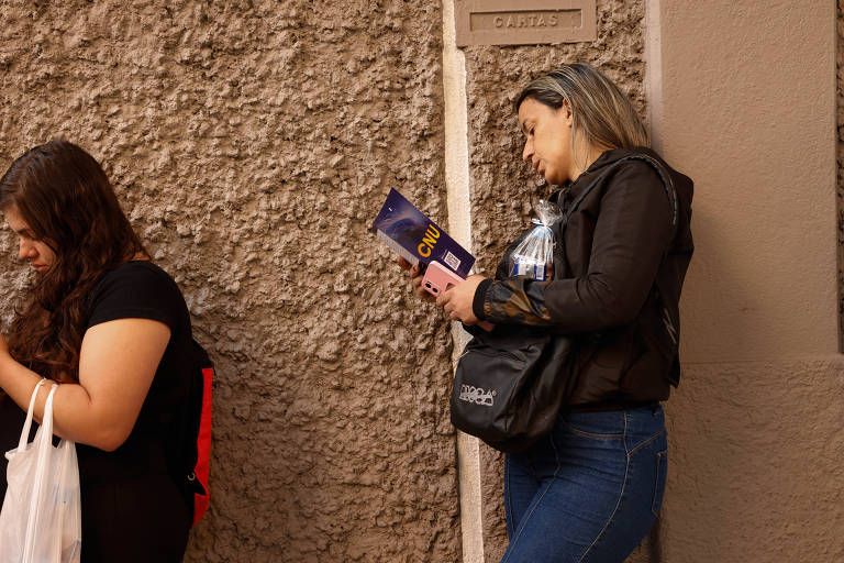 A imagem mostra duas mulheres em um ambiente urbano. À esquerda, uma mulher com cabelo longo e escuro, vestindo uma camiseta preta, está olhando para o celular. À direita, uma mulher com cabelo longo e claro, vestindo uma jaqueta preta e jeans, está encostada em uma parede e segurando um folheto. O fundo é uma parede texturizada de cor clara.