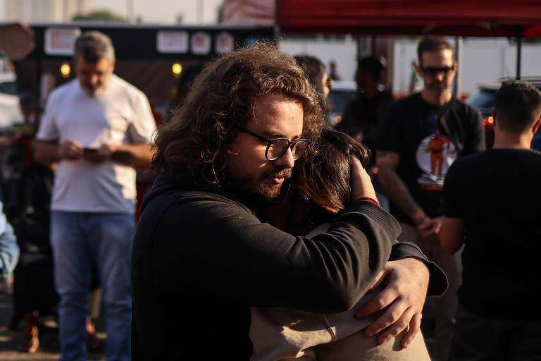 A imagem mostra um homem com cabelo cacheado e óculos， abraçando uma mulher. Eles parecem estar em um ambiente ao ar livre， com várias pessoas ao fundo， algumas olhando para seus celulares. A luz do sol ilumina a cena， criando um clima de afeto e conexão entre os dois.