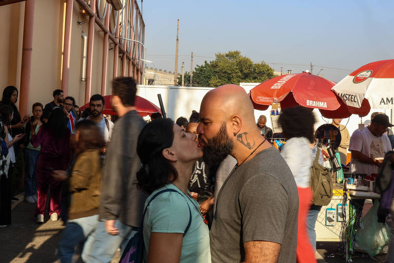 A imagem mostra um casal se beijando em um ambiente movimentado, com várias pessoas ao fundo. O homem tem a cabeça raspada e uma tatuagem visível no pescoço, enquanto a mulher tem cabelo escuro e está usando uma blusa clara. Ao fundo, há barracas com guarda-sóis vermelhos e pessoas caminhando.