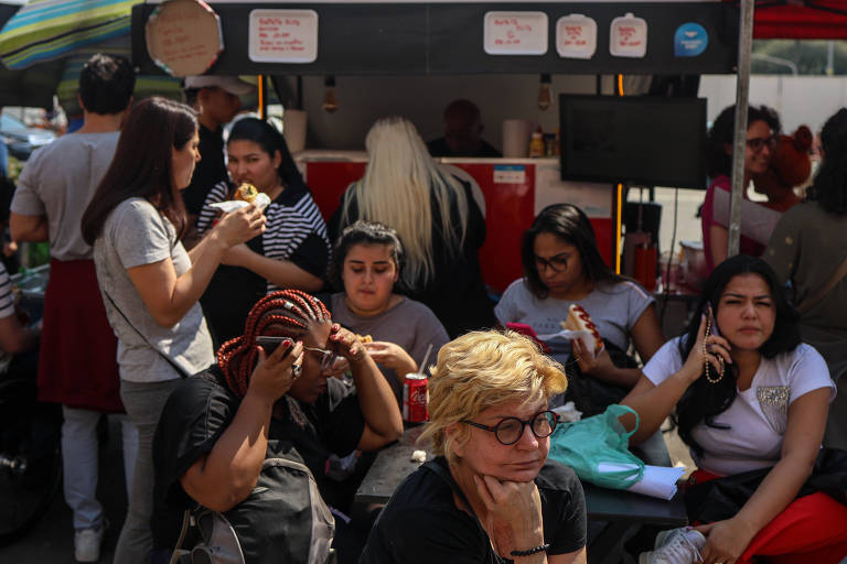 A imagem mostra um grupo de pessoas em um ambiente de feira de comida de rua. Algumas pessoas estão em pé， segurando alimentos， enquanto outras estão sentadas. Ao fundo， há um quiosque de comida com uma tela e placas visíveis. A cena é movimentada， com várias pessoas interagindo entre si.
