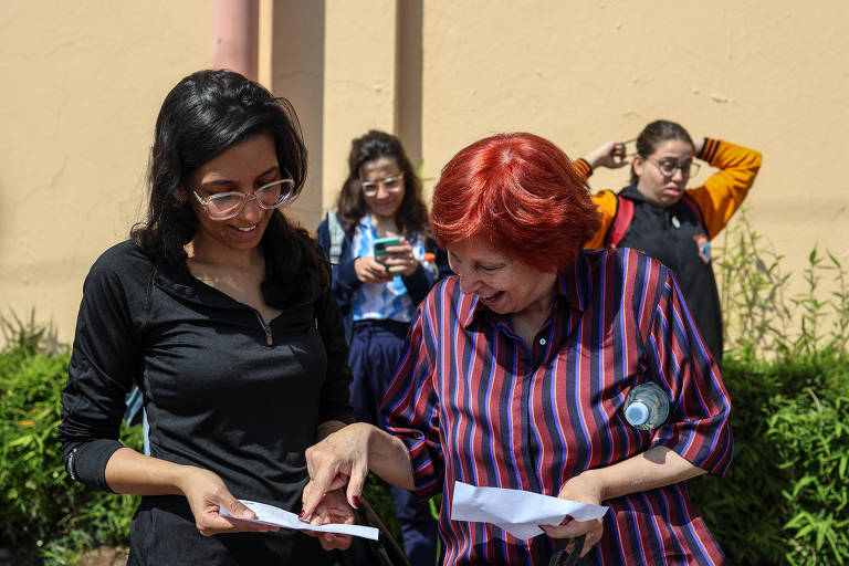 A imagem mostra duas mulheres em primeiro plano， uma com cabelo longo e escuro， usando óculos， e a outra com cabelo curto e vermelho， vestindo uma camisa listrada. Elas estão olhando para papéis que seguram nas mãos e parecem estar conversando. Ao fundo， há duas outras mulheres， uma com cabelo preso e outra com uma blusa laranja， ambas observando a cena.