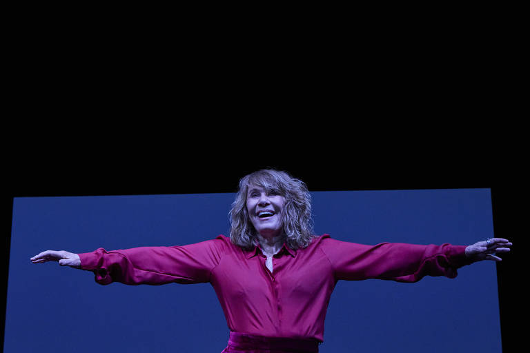 Uma mulher com cabelo cacheado e loiro, vestindo uma camisa vermelha, está em um palco com os braços abertos, sorrindo. O fundo é escuro, com uma tela azul clara atrás dela