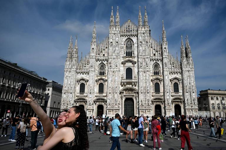 Imagem mostra o Duomo de Milão, uma imponente estrutura gótica com várias torres e detalhes elaborados. Em primeiro plano, duas pessoas estão tirando uma selfie, enquanto um grupo de pessoas caminha na praça em frente à catedral. O céu está parcialmente nublado, e a catedral é o foco central da imagem.