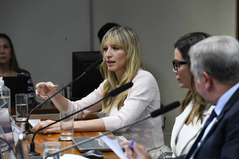 Mulher loira de blusa branca falando ao microfone na sala da comissão