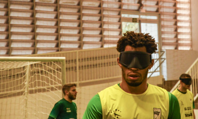 Foto de Karime Xavier mostra o busto de Maicon Júnior durante um treino da seleção. Ele é um Homem Negro retinto, com cabelos nudreads com luzes. Ele veste o colete amarelo da seleção por cima da camisa verde de treino. 