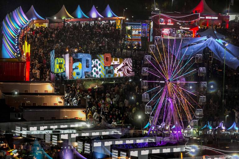 A imagem mostra um festival noturno com uma grande multidão. Ao fundo, há várias tendas coloridas e uma roda gigante iluminada com luzes coloridas. Em destaque, há uma estrutura com letras grandes e coloridas formando a palavra 'LOLLAPALOOZA'. O ambiente é vibrante, com muitas luzes e uma atmosfera festiva.