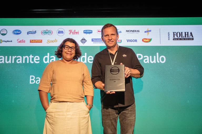 A imagem mostra duas pessoas em um evento de premiação. À esquerda, uma mulher de óculos, vestindo uma blusa clara e calças brancas. À direita, um homem com cabelo curto, usando uma camisa escura e segurando um prêmio. Ao fundo, uma tela verde com logotipos de várias marcas.