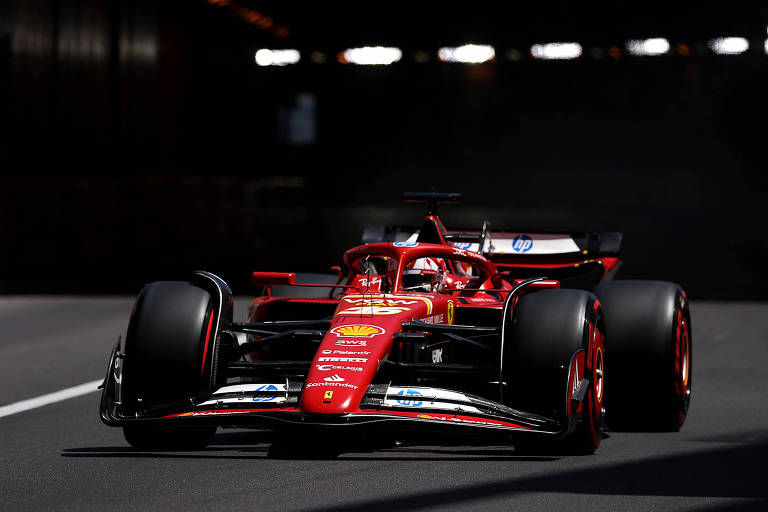 Um carro de Fórmula 1, da Ferrari, predominantemente vermelho, está em movimento em um túnel. O veículo apresenta detalhes aerodinâmicos e rodas largas, com luzes refletindo no ambiente escuro do túnel.
