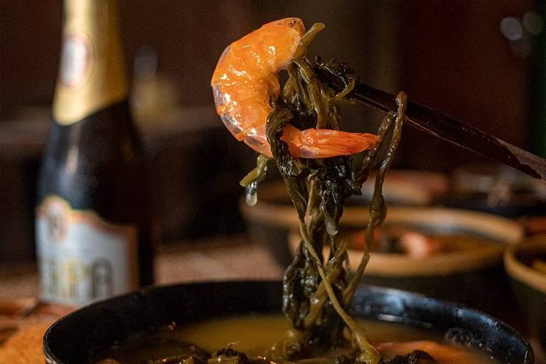 A imagem mostra um prato de comida típica, com camarões e folhas verdes em um caldo. Um camarão é segurado por um par de hashis, enquanto o prato é decorado com a palavra 'Paraense'. Ao fundo, há uma garrafa de cerveja e outros pratos.