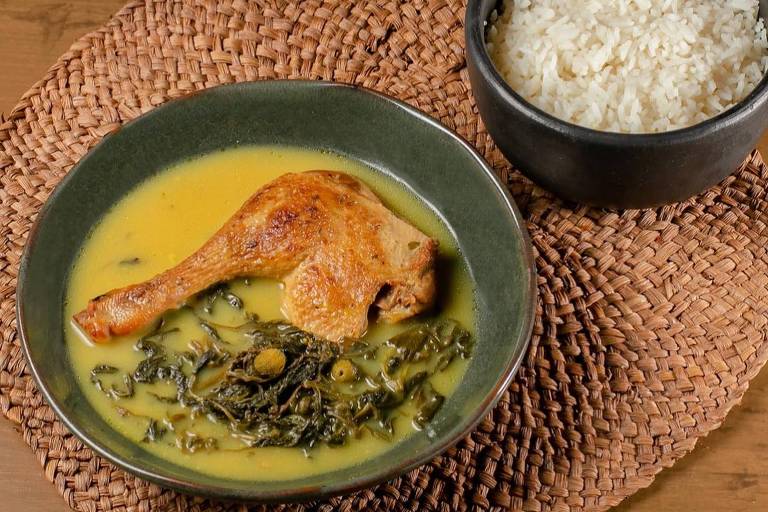 A imagem mostra um prato de pato com molho amarelo e folhas verdes, acompanhado de arroz branco em uma tigela. O prato está sobre um suporte de palha, e o fundo é uma mesa de madeira.