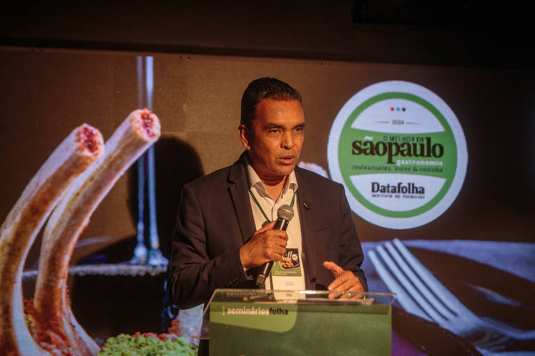 Um homem em um terno escuro está falando ao microfone em um evento. Ao fundo, há uma imagem de pratos de comida, incluindo costelas e vegetais. Um logotipo com as palavras 'São Paulo' e 'Datafolha' é visível na parte superior direita da imagem.