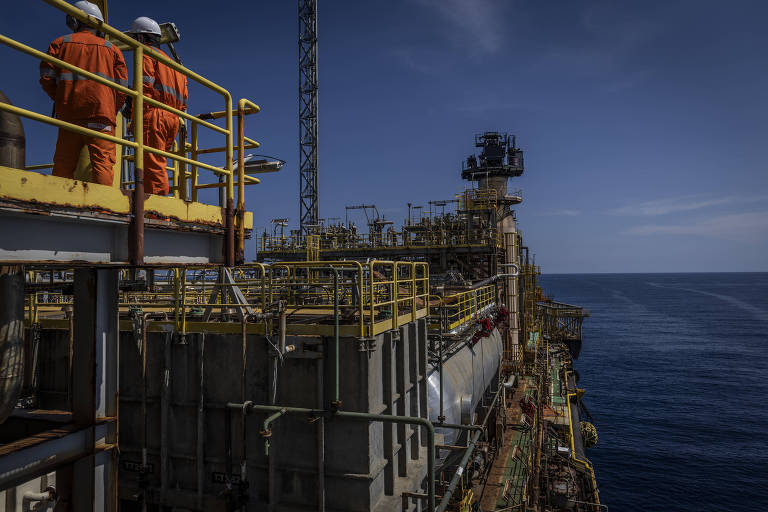 A imagem mostra uma plataforma de petróleo no mar, com dois trabalhadores vestidos com macacões laranjas observando o horizonte. A estrutura da plataforma é composta por várias tubulações e equipamentos, e o céu está claro, com algumas nuvens. O mar é visível ao fundo, refletindo a luz do dia.