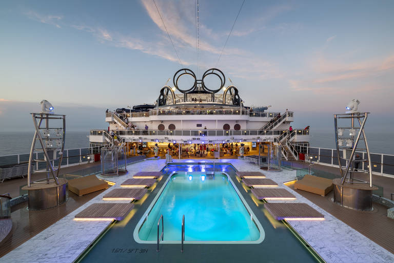 A imagem mostra a área da piscina em um navio de cruzeiro， com uma piscina iluminada em azul e espreguiçadeiras ao redor. Ao fundo， há um bar e várias pessoas se divertindo. O céu está claro， com algumas nuvens， e o mar é visível ao fundo.