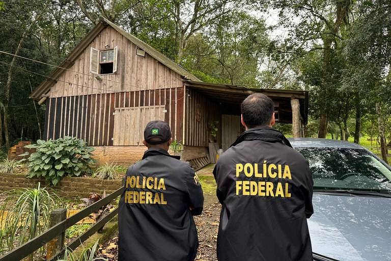 A imagem mostra dois agentes da Polícia Federal, de costas, observando uma casa de madeira em uma área arborizada. Os agentes estão vestidos com jaquetas pretas que têm a inscrição 'POLÍCIA FEDERAL' em letras amarelas. Ao fundo, a casa apresenta uma estrutura simples, com janelas e uma porta fechada, cercada por vegetação