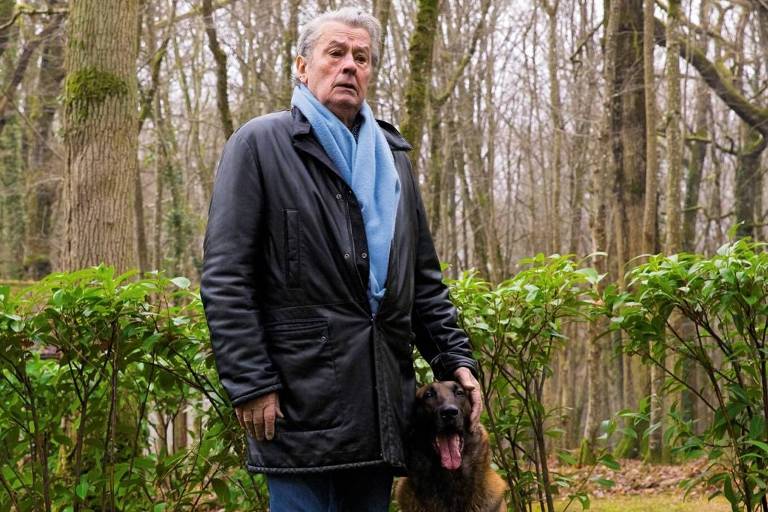 Um homem de cabelos grisalhos, vestindo um casaco preto e um cachecol azul, está em pé ao lado de um cão da raça Malinois. O homem parece sério e está em uma área florestal com árvores ao fundo e vegetação verde ao redor. O cão está sentado ao lado dele, com a boca aberta, parecendo relaxado. O chão é coberto por musgo.