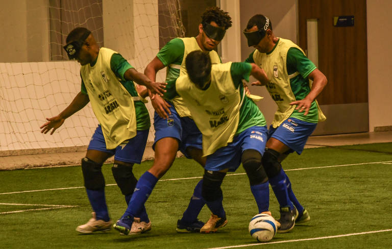 Foto de Karime Xavier mostra quatro jogadores da seleção de futebol de cegos do Brasil. Eles estão vestindo chuteiras, meiões e calções azuis, camisa verde e um colete amarelo por cima. Todos estão usando vendas pretas