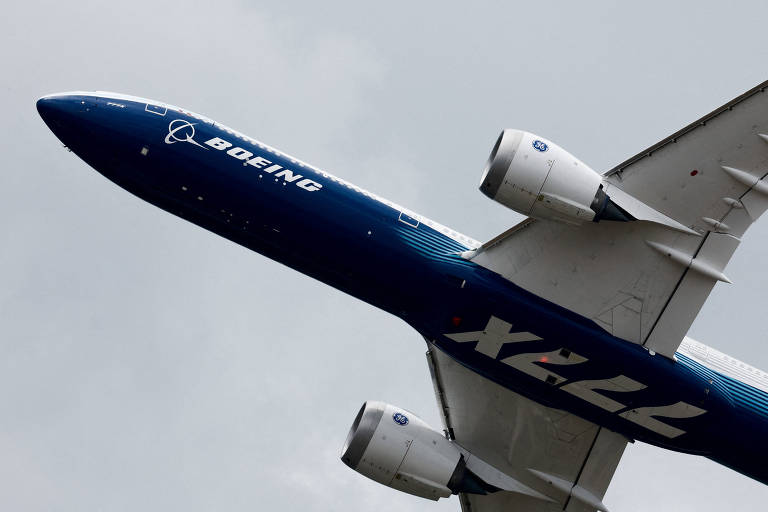 A imagem mostra um avião Boeing em um ângulo inclinado, com a fuselagem predominantemente azul e a inscrição 'BOEING' em letras brancas. O número 'X22' também é visível na parte inferior do avião. O fundo é nublado, sugerindo que o avião está voando em um céu encoberto.
