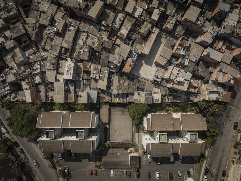 Imagem aérea mostra dois telhados grandes, que são de prédios, com centenas de talhados de casas próximos, tudo em tons de cinza