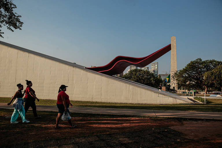 A imagem mostra três pessoas caminhando em uma área externa, com um grande edifício moderno ao fundo. O edifício possui uma estrutura inclinada e uma cobertura vermelha ondulada, contrastando com a parede branca. À direita, há um monumento em forma de obelisco. O céu está claro, indicando um dia ensolarado.