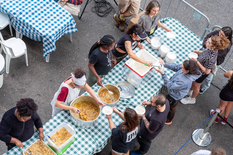 A imagem mostra uma cena de distribuição de alimentos em um evento comunitário. Várias pessoas estão envolvidas no processo, algumas servindo comida de grandes panelas e outras recebendo porções em pratos. As mesas estão cobertas com toalhas de mesa quadriculadas em azul e branco. Há uma variedade de pratos, incluindo um grande recipiente com um guisado e outro com arroz. O ambiente parece ser ao ar livre, com cadeiras brancas e uma barreira de segurança ao fundo.